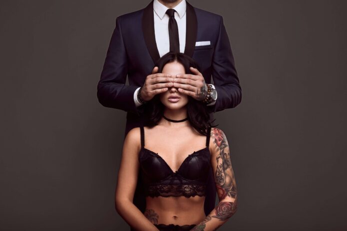 portrait-of-businessman-in-elegant-suit-cover-eyes-of-sexy-woman-with-tattoo-in-lingerie-min (1)