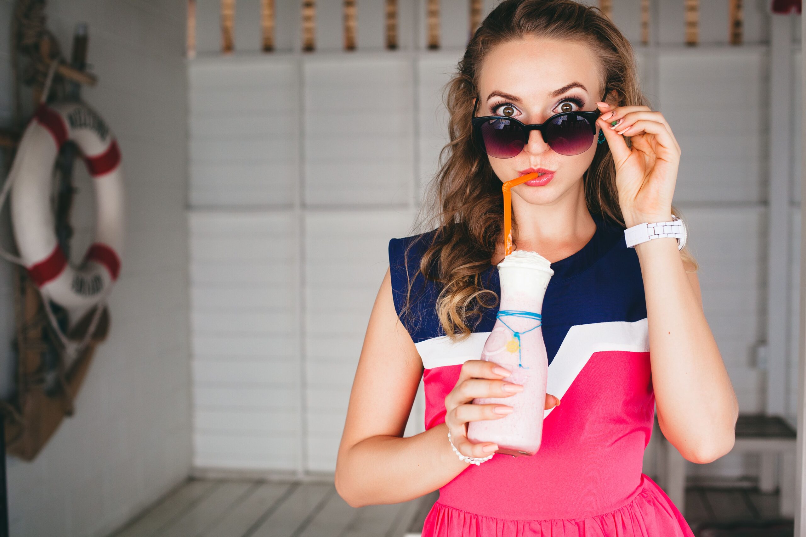 young-stylish-beautiful-woman-in-sea-cafe-drinking-cocktail-smoothie-sunglasses-flirty-resort-style-fashionable-outfit-smiling-marine-colors-dress-anchor-and-lifebuoy-on-background-shocked-min
