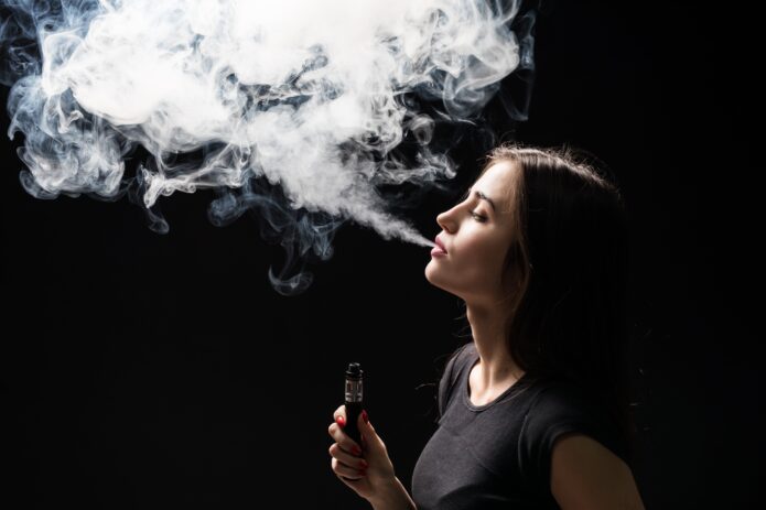 young-beautiful-brunette-woman-smoking-vaping-e-cigarette-with-smoke-on-the-black-wall-min